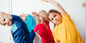 Young children stretching with arms overhead during a group exercise activity indoors, wearing colorful shirts and following along together