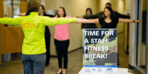 Group of employees participating in a workplace fitness break with arms outstretched in a hallway next to a sign reading &ldquo;Time for a Staff Fitness Break!&rdquo;